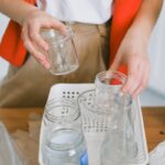 Crop anonymous female in casual clothes putting glass jars and bottles into plastic container while sorting waste in kitchen